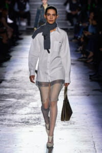 Model walks the runway in an oversized white button-down shirt, sheer lace shorts, and a gray knit scarf, carrying a brown tote bag.
