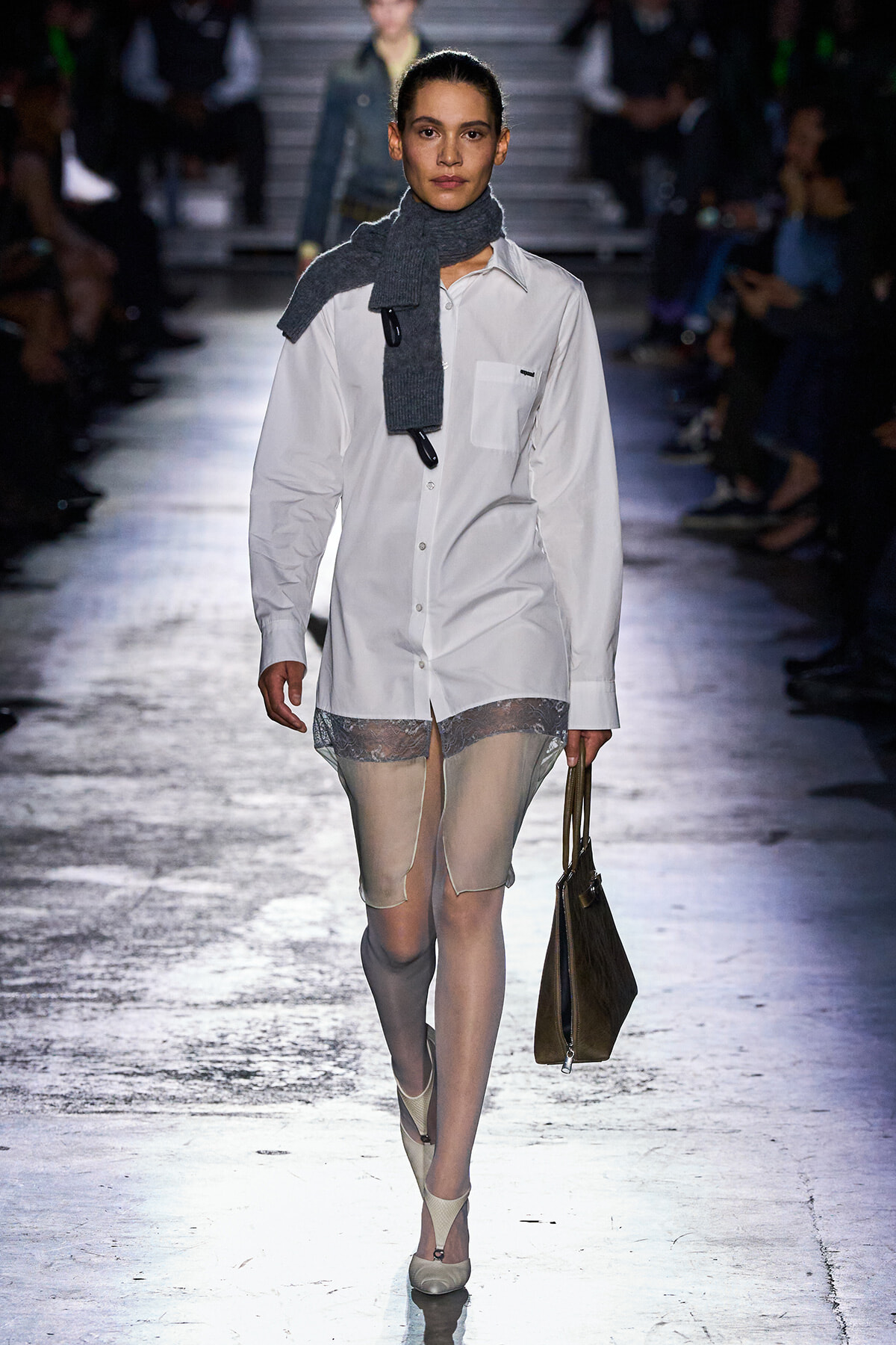 Model walks the runway in an oversized white button-down shirt, sheer lace shorts, and a gray knit scarf, carrying a brown tote bag.