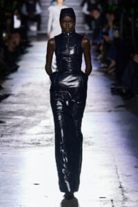 Model walks a fashion show in a dark, glossy padded jumpsuit with a hooded headpiece .