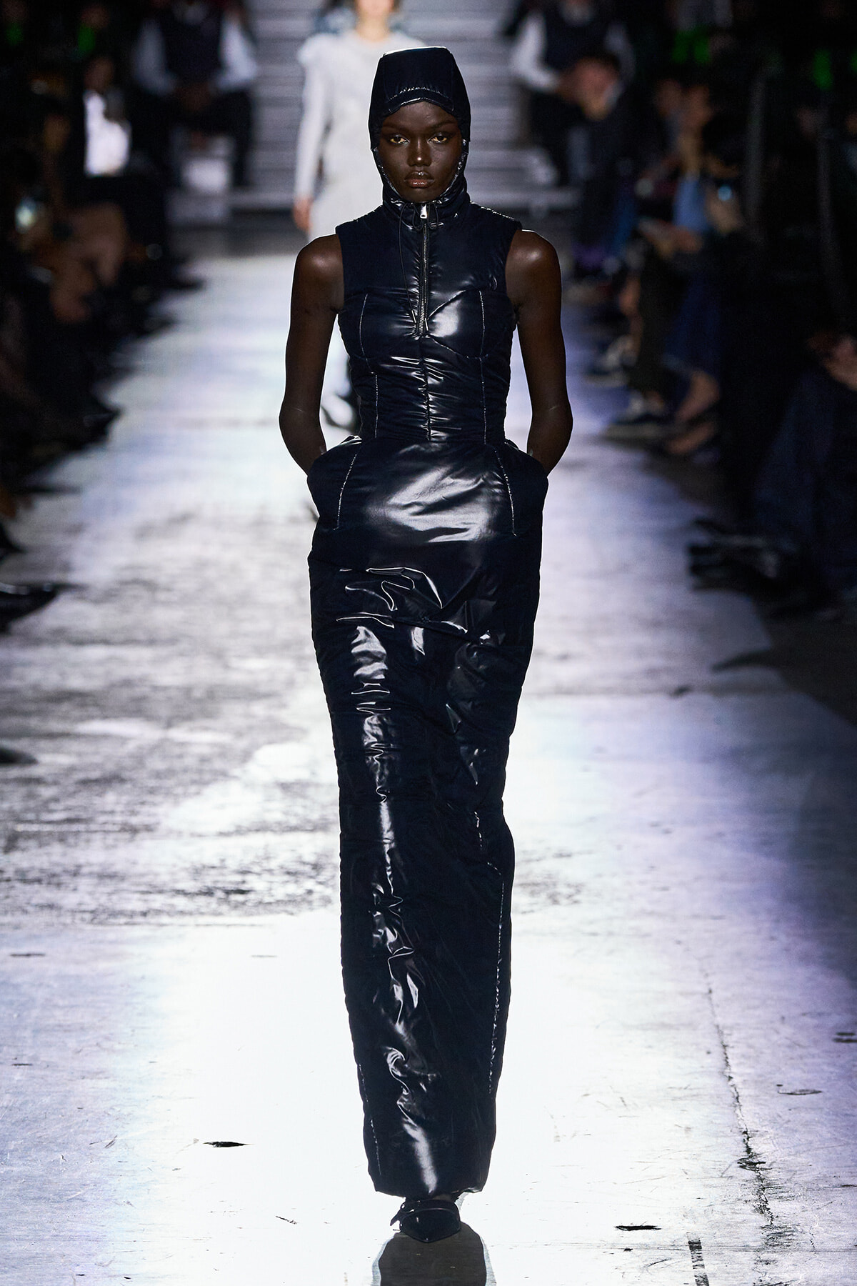 Model walks a fashion show in a dark, glossy padded jumpsuit with a hooded headpiece .