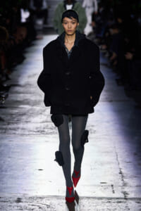 Model walking a runway in a black oversized coat, gray ribbed tights, and bright red pointed heels.