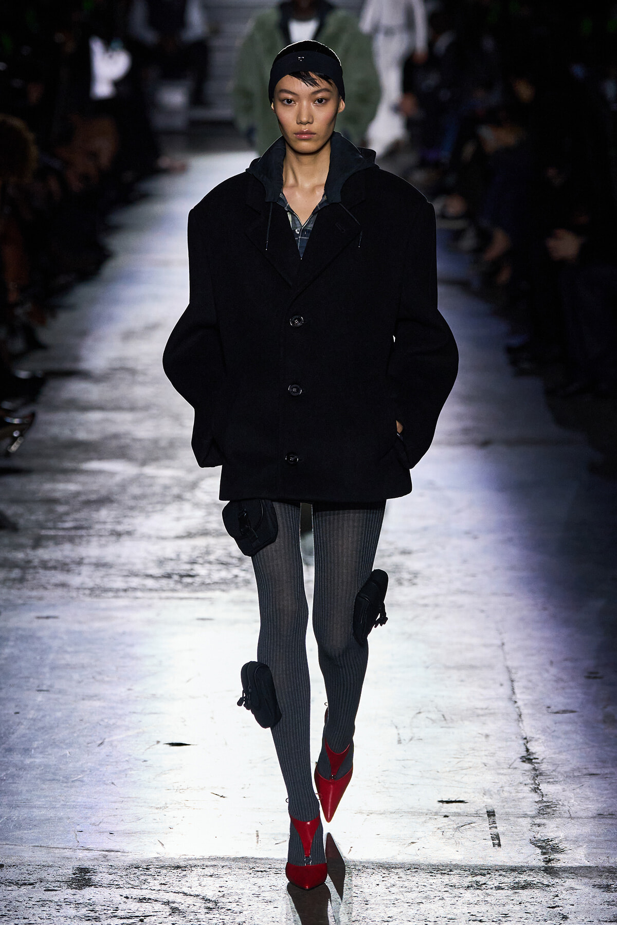 Model walking a runway in a black oversized coat, gray ribbed tights, and bright red pointed heels.