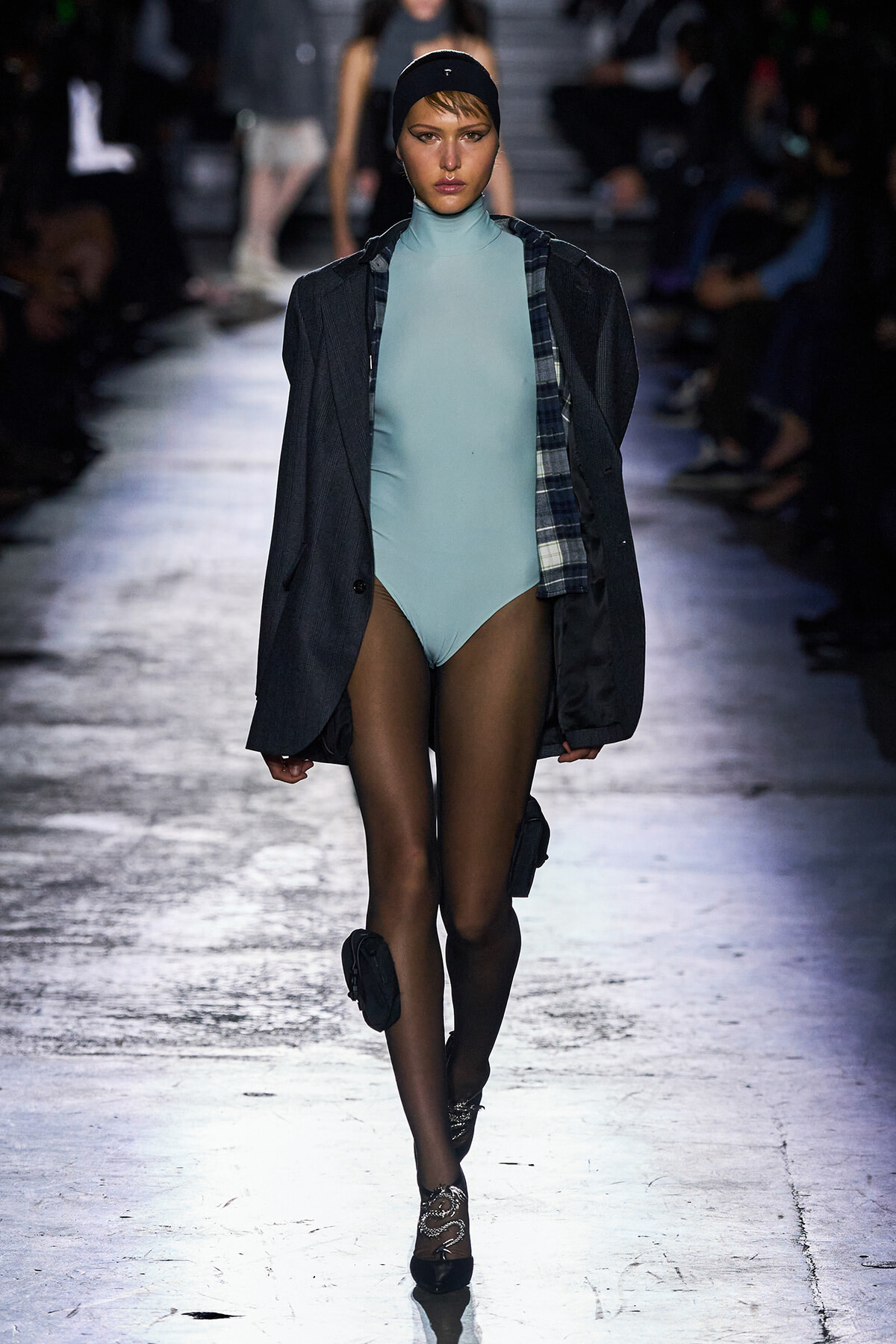 Model on a runway wearing a light blue high-neck bodysuit under a dark oversized blazer with a plaid shirt, black tights, and black heels with ankle accessories.