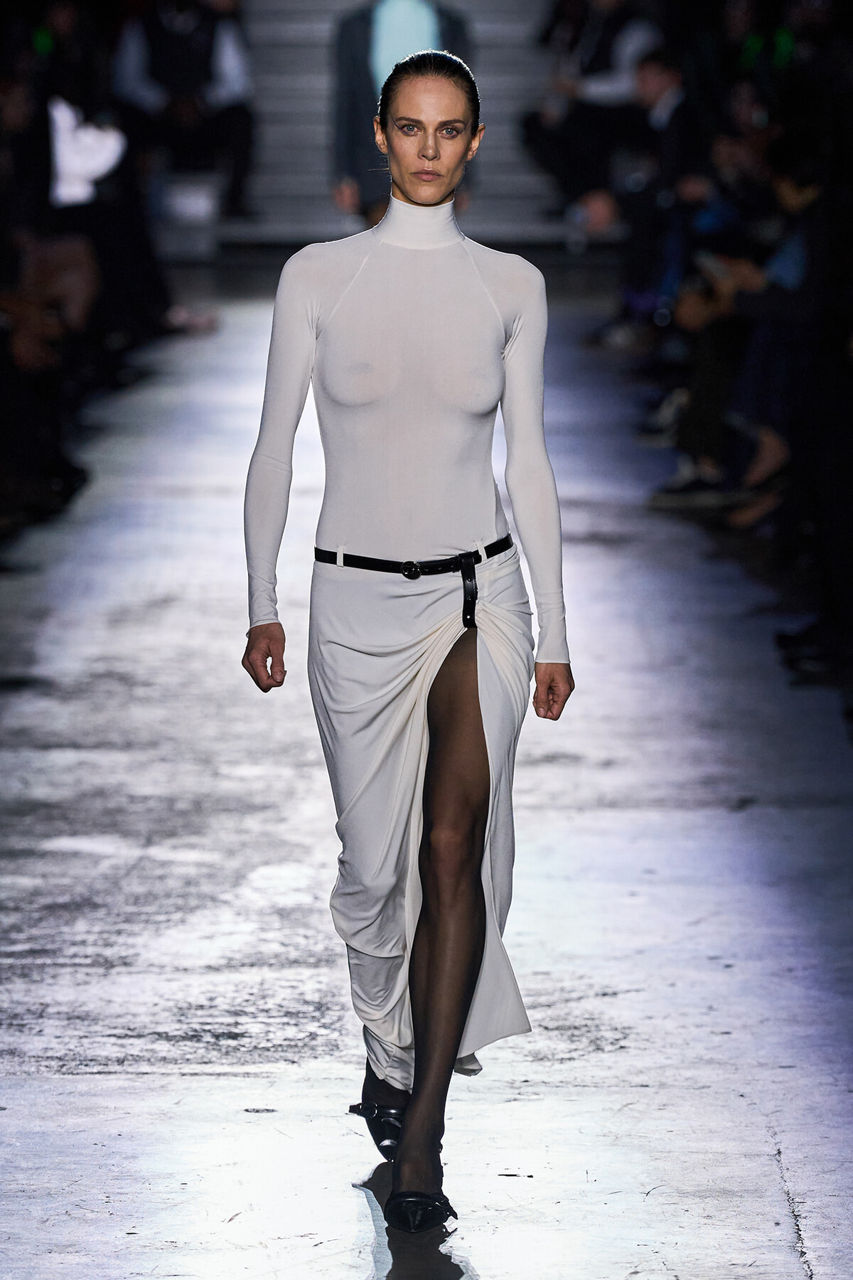 Model walking a runway in a white turtleneck bodysuit and a high-slit white skirt with a black belt.