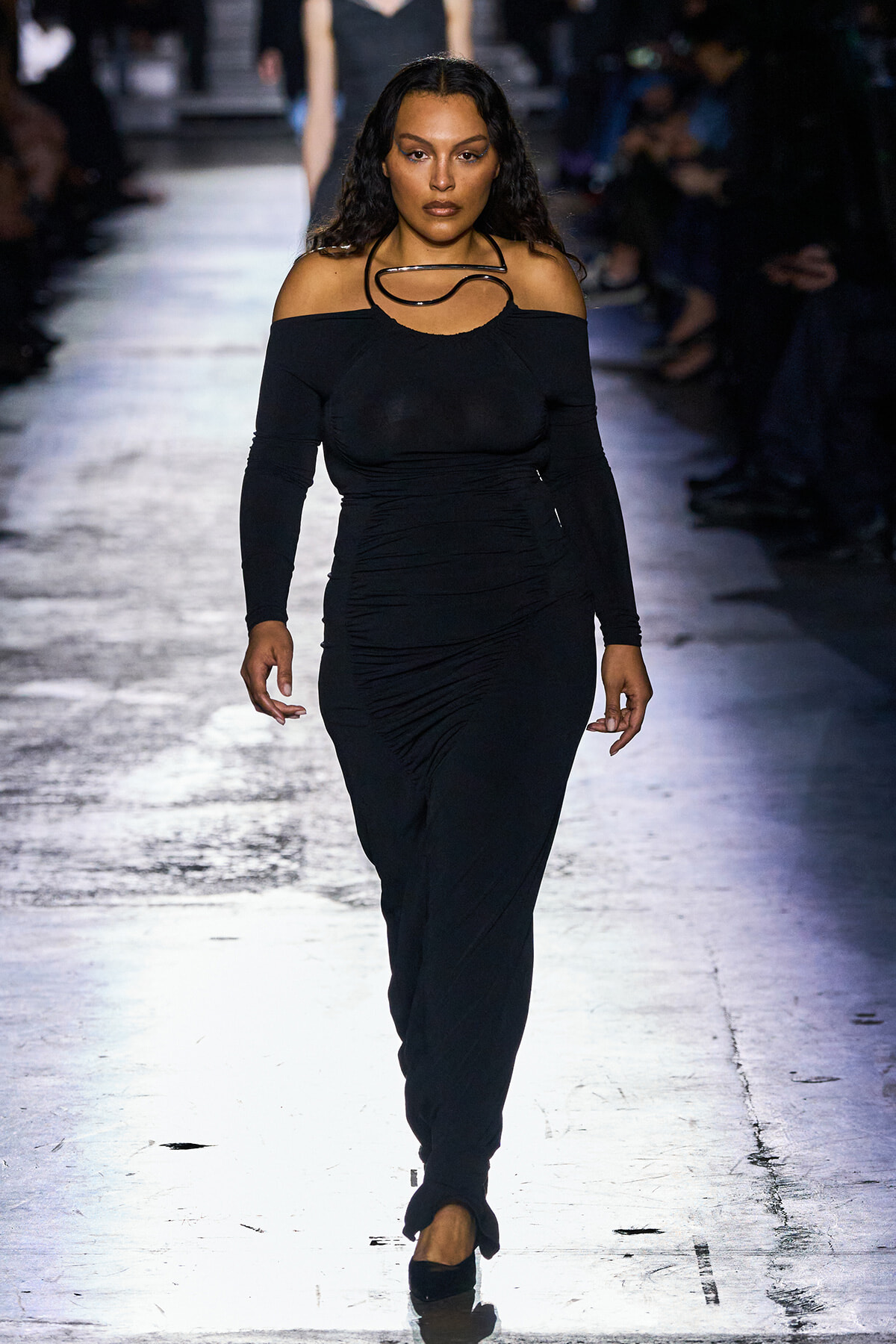 Model walks the runway in a black off-the-shoulder, long-sleeve dress with a large circular necklace; spotlighted catwalk with a blurred audience in the background.