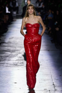 Model walks the runway in a strapless, red shiny jumpsuit with pockets at the hips.