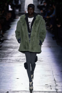Runway model in an oversized olive-green faux-fur coat over a plaid shirt, gray ribbed tights, and beige heels.