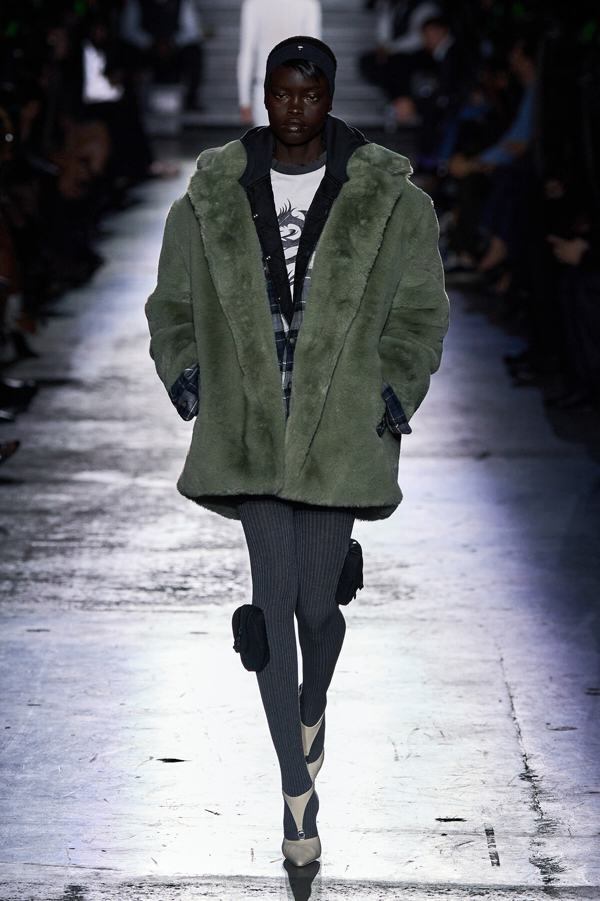 Runway model in an oversized olive-green faux-fur coat over a plaid shirt, gray ribbed tights, and beige heels.