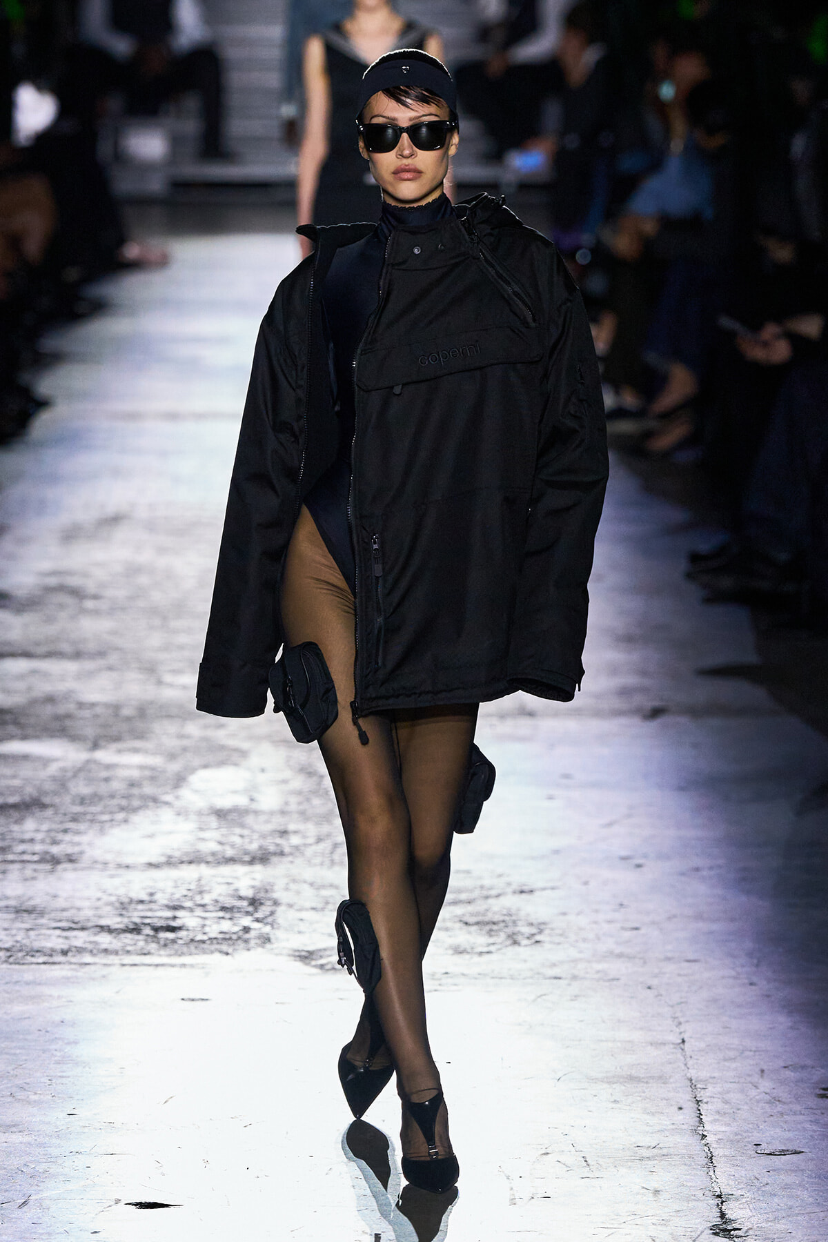 Female model walking a fashion runway in a bulky black jacket, black cap, oversized sunglasses, sheer tights, and black heels with small accessory pouches.