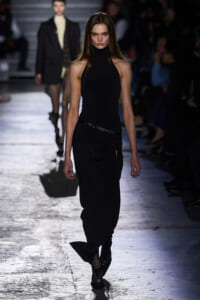 Model walks the runway in a black sleeveless turtleneck top and draped black skirt with a belt.