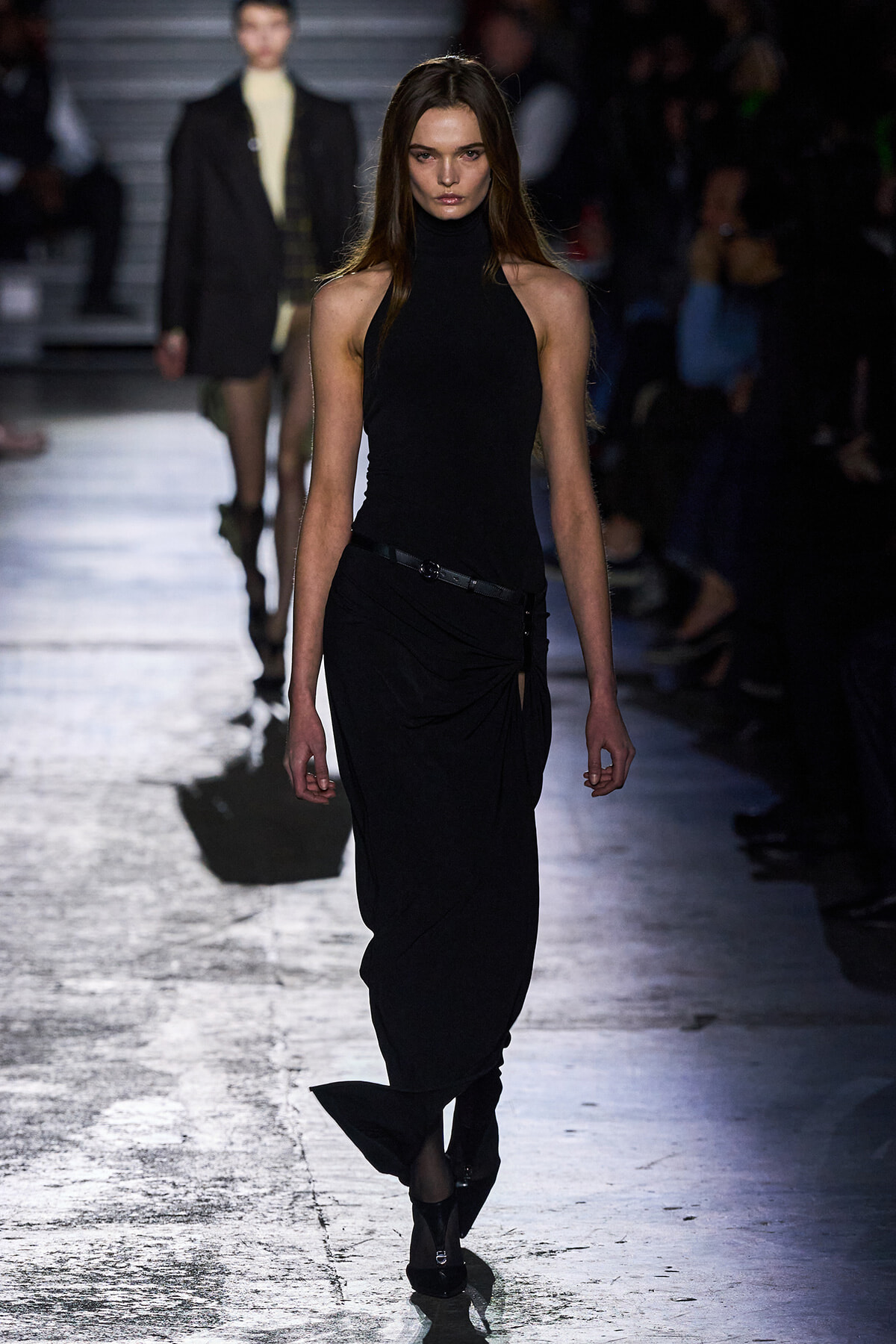 Model walks the runway in a black sleeveless turtleneck top and draped black skirt with a belt.