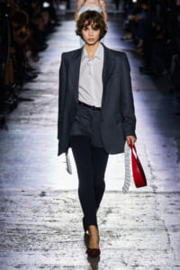 Model walking runway in oversized gray suit with white shirt, black tights, and pointed burgundy heels, carrying a red tote with white fringe.