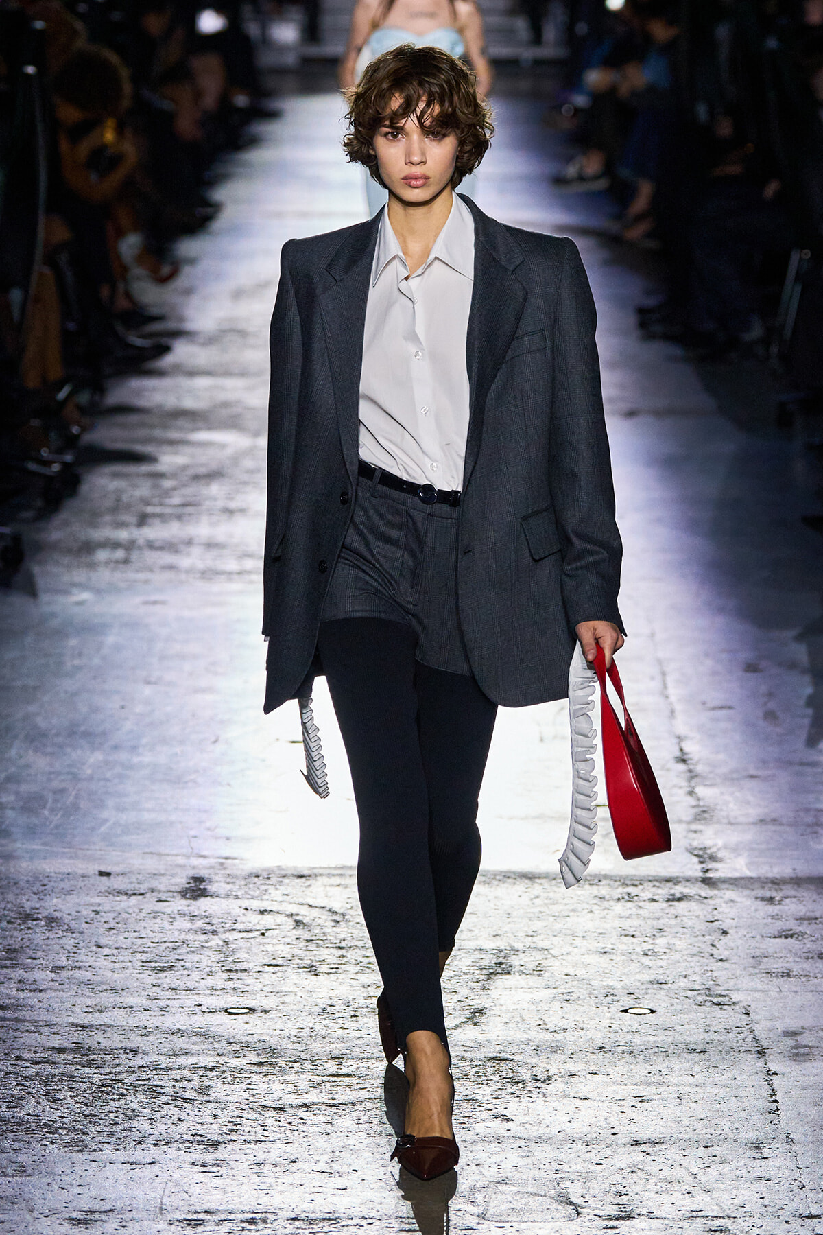 Model walking runway in oversized gray suit with white shirt, black tights, and pointed burgundy heels, carrying a red tote with white fringe.