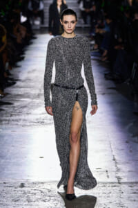 Model walks down a fashion runway in a long speckled grey knit dress with a high thigh slit and black belt.