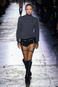 Runway model walking in a gray turtleneck sweater with black studded harness shorts and knee-high black boots.