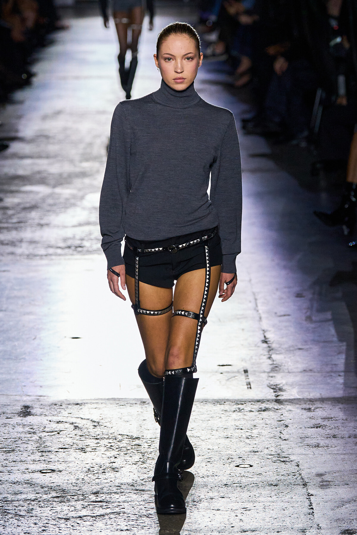 Runway model walking in a gray turtleneck sweater with black studded harness shorts and knee-high black boots.