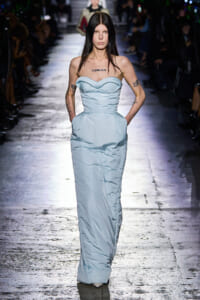 Model walking a fashion show wearing a pale blue strapless gown with a structured bodice and pockets, tattoos visible on arms and a 'GENESIS' tattoo on the chest.