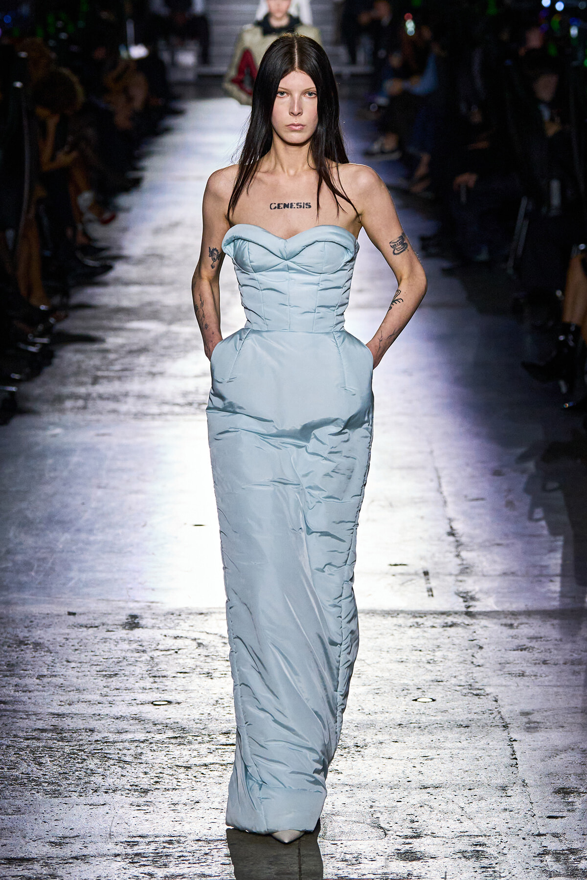 Model walking a fashion show wearing a pale blue strapless gown with a structured bodice and pockets, tattoos visible on arms and a 'GENESIS' tattoo on the chest.