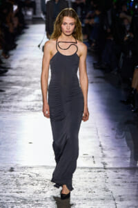 Model walks a fashion runway in a sleeveless, ruched, charcoal-gray gown with a sculptural black necklace.