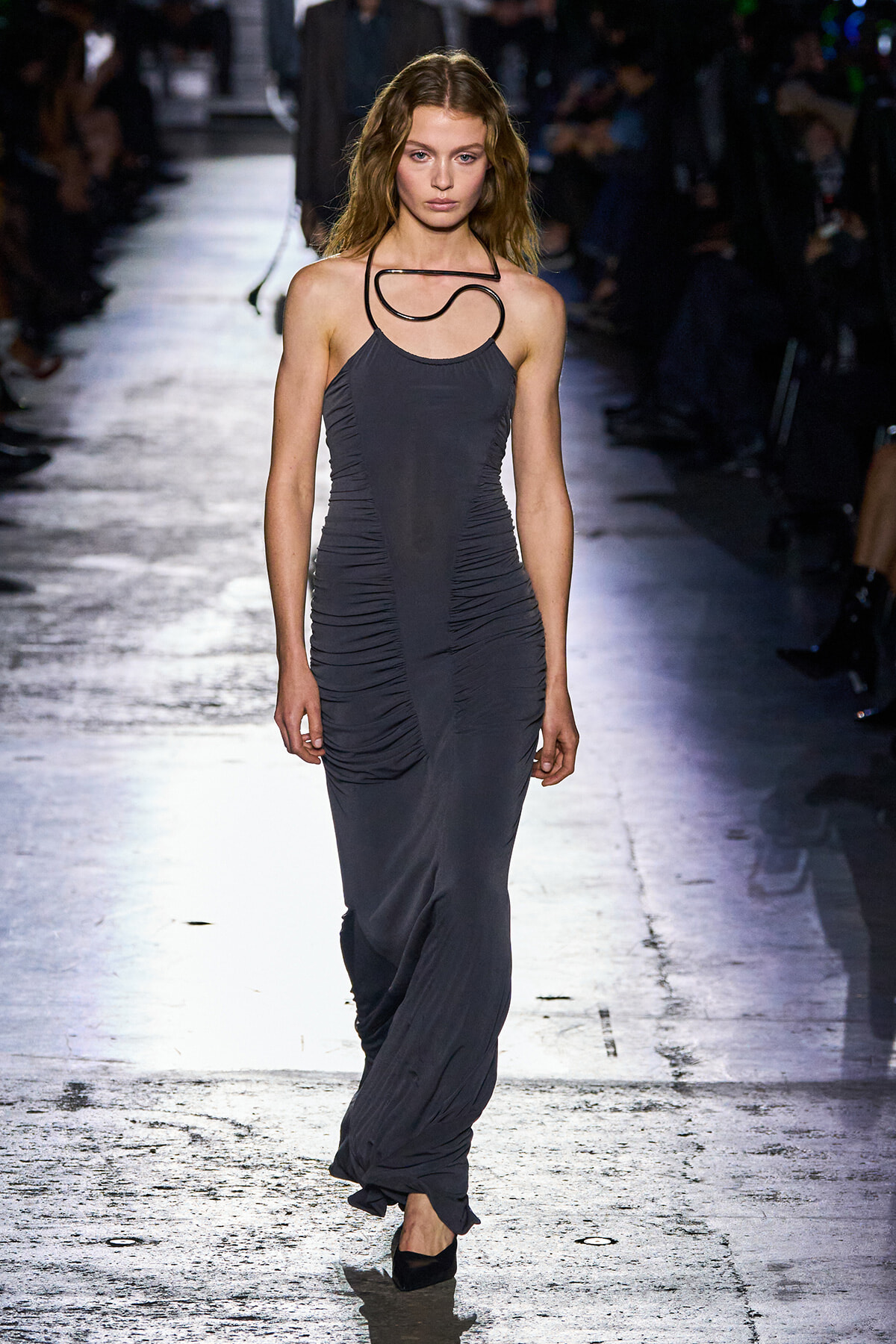 Model walks a fashion runway in a sleeveless, ruched, charcoal-gray gown with a sculptural black necklace.