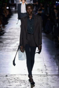 Model walks a runway in a dark pinstriped suit with a black shirt, holding long black spiral straps in both hands.