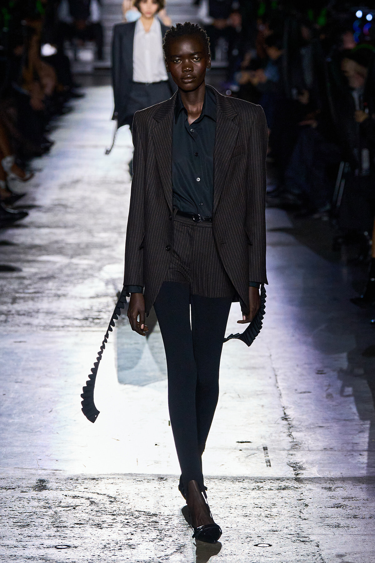 Model walks a runway in a dark pinstriped suit with a black shirt, holding long black spiral straps in both hands.