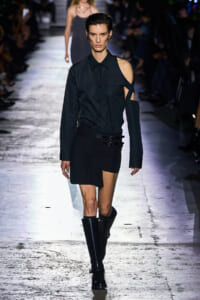 Runway model in an all-black, asymmetric outfit with a one-shoulder cut and knee-high boots on a dimly lit catwalk.