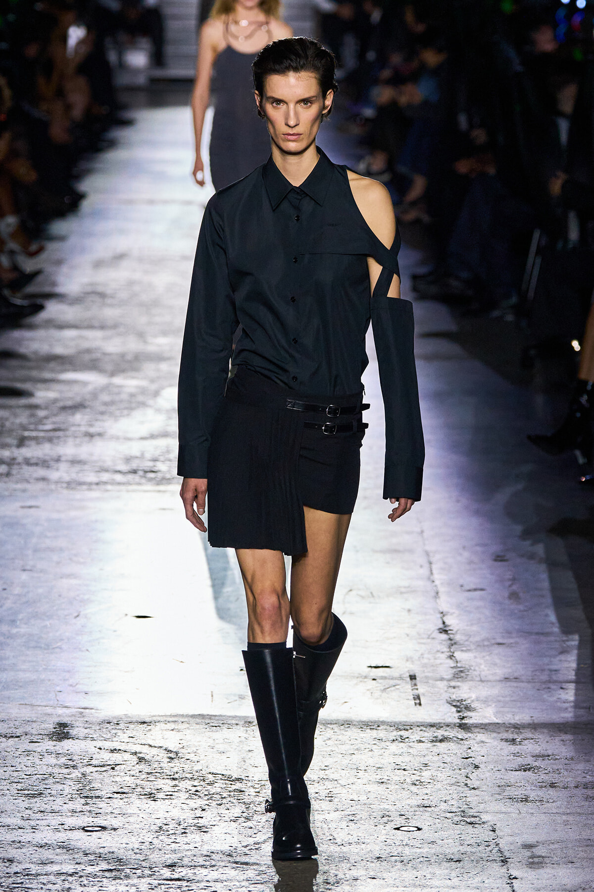 Runway model in an all-black, asymmetric outfit with a one-shoulder cut and knee-high boots on a dimly lit catwalk.