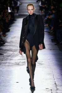 Runway model walking in a black blazer over a dark bodysuit with sheer tights and small accessories clipped to her legs, audience in the background.
