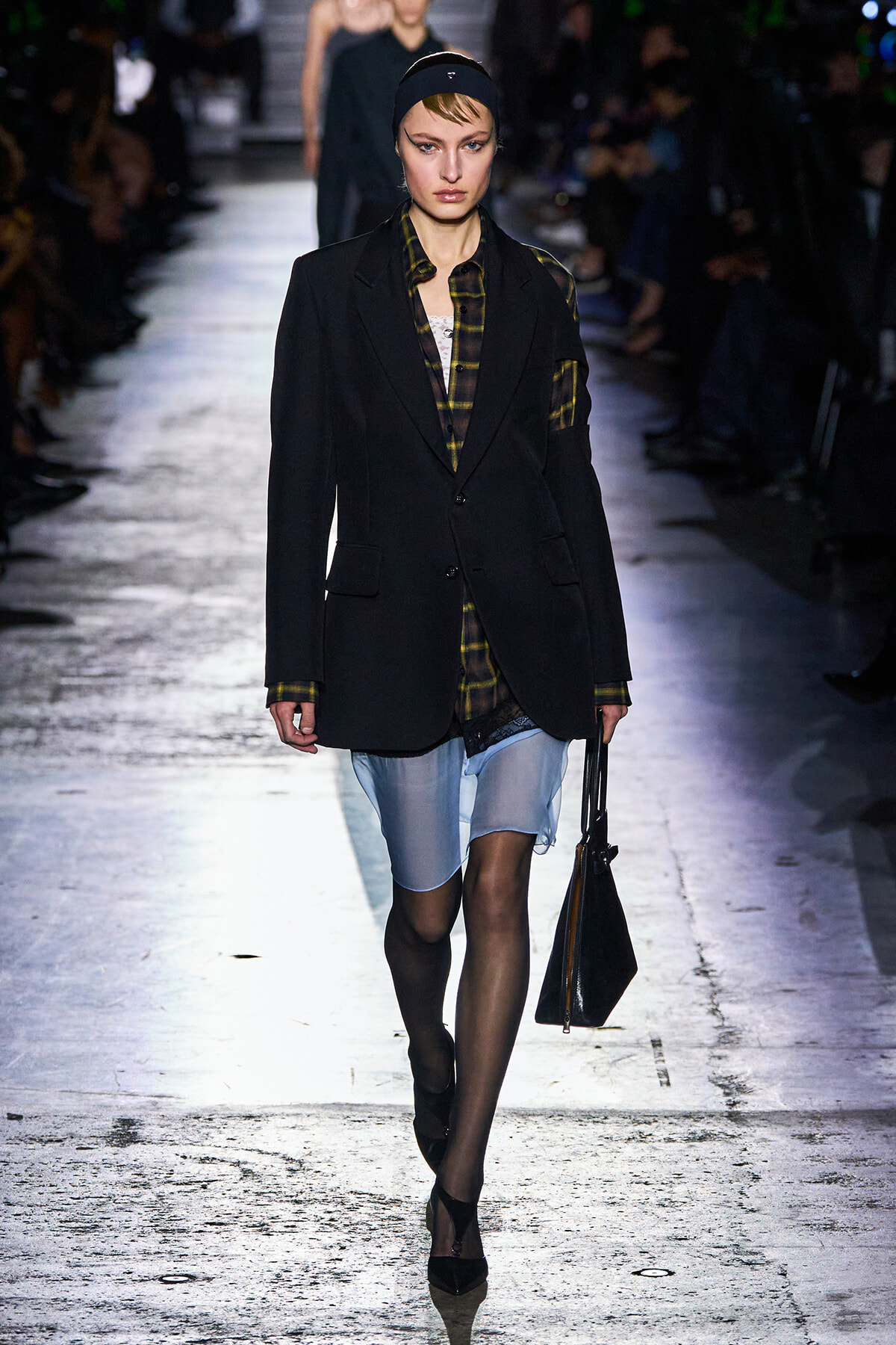 Model walking a fashion runway in a black blazer over a plaid shirt, with light denim shorts and a black handbag.