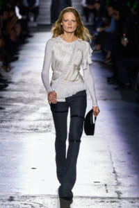 Model walking a runway in a white pleated blouse and dark jeans, holding a small black hat at her side.