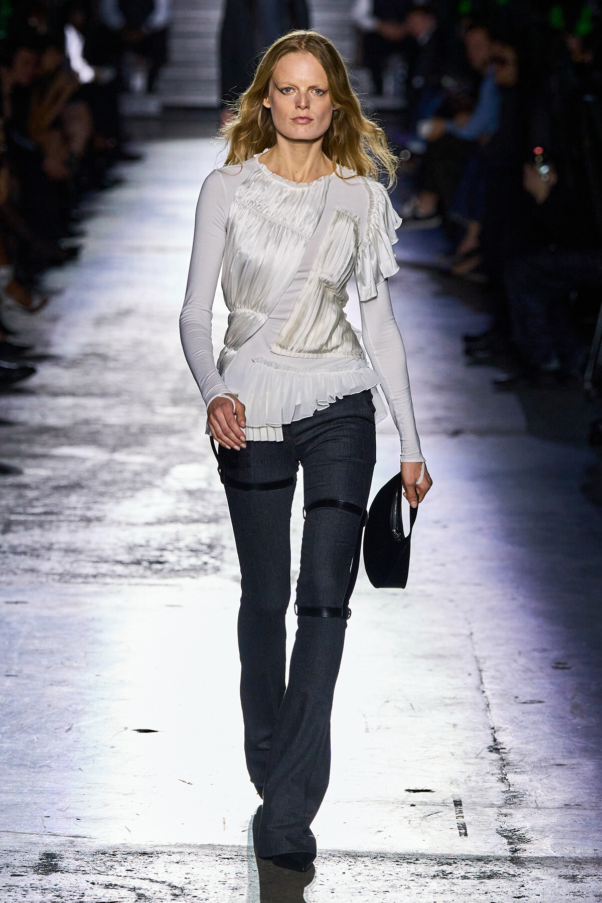 Model walking a runway in a white pleated blouse and dark jeans, holding a small black hat at her side.
