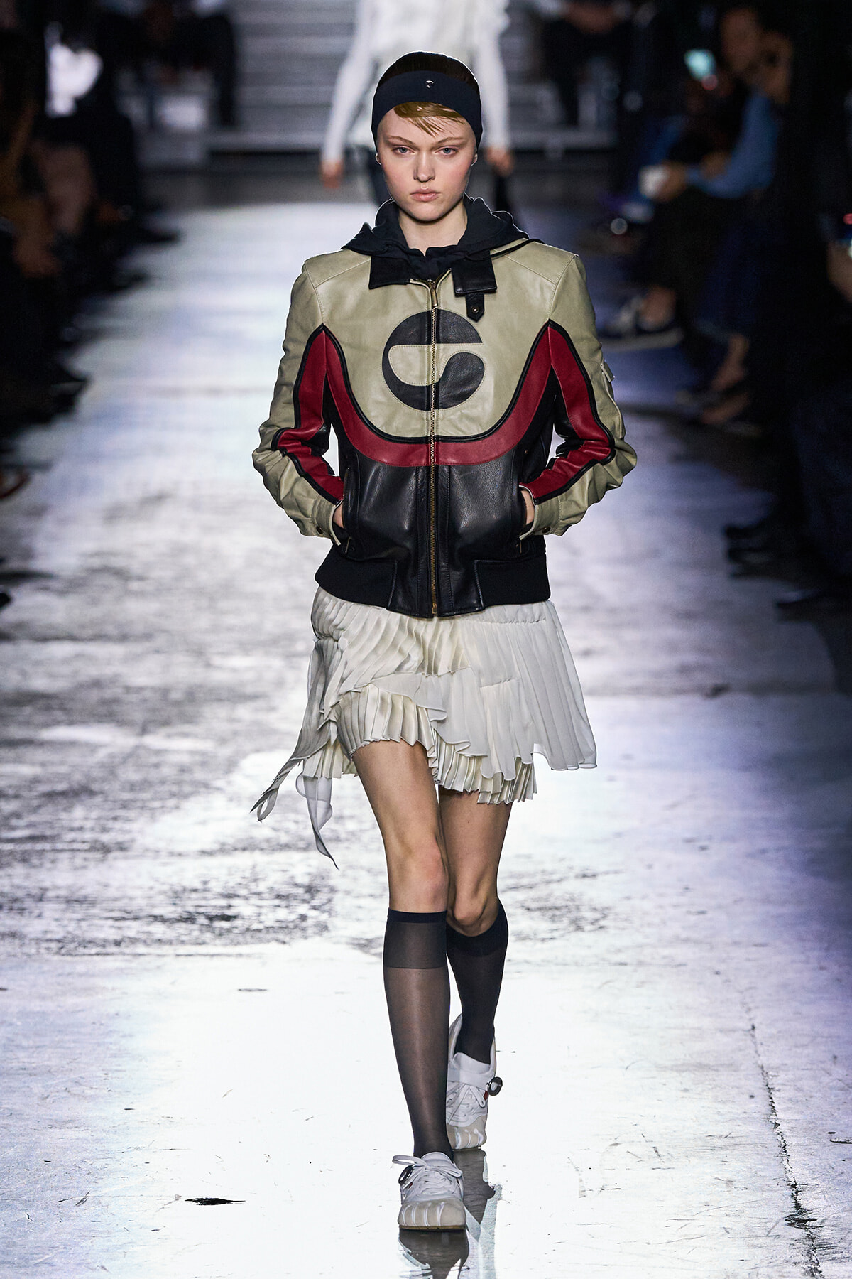 Model on runway wearing a beige, black, and red color-block leather bomber with a white pleated skirt and headband beneath; hands in pockets.