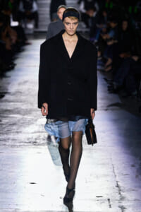 Model walks a fashion show in an oversized black coat over a light blue sheer dress, black tights, headband, and carrying a black handbag on a dim runway.