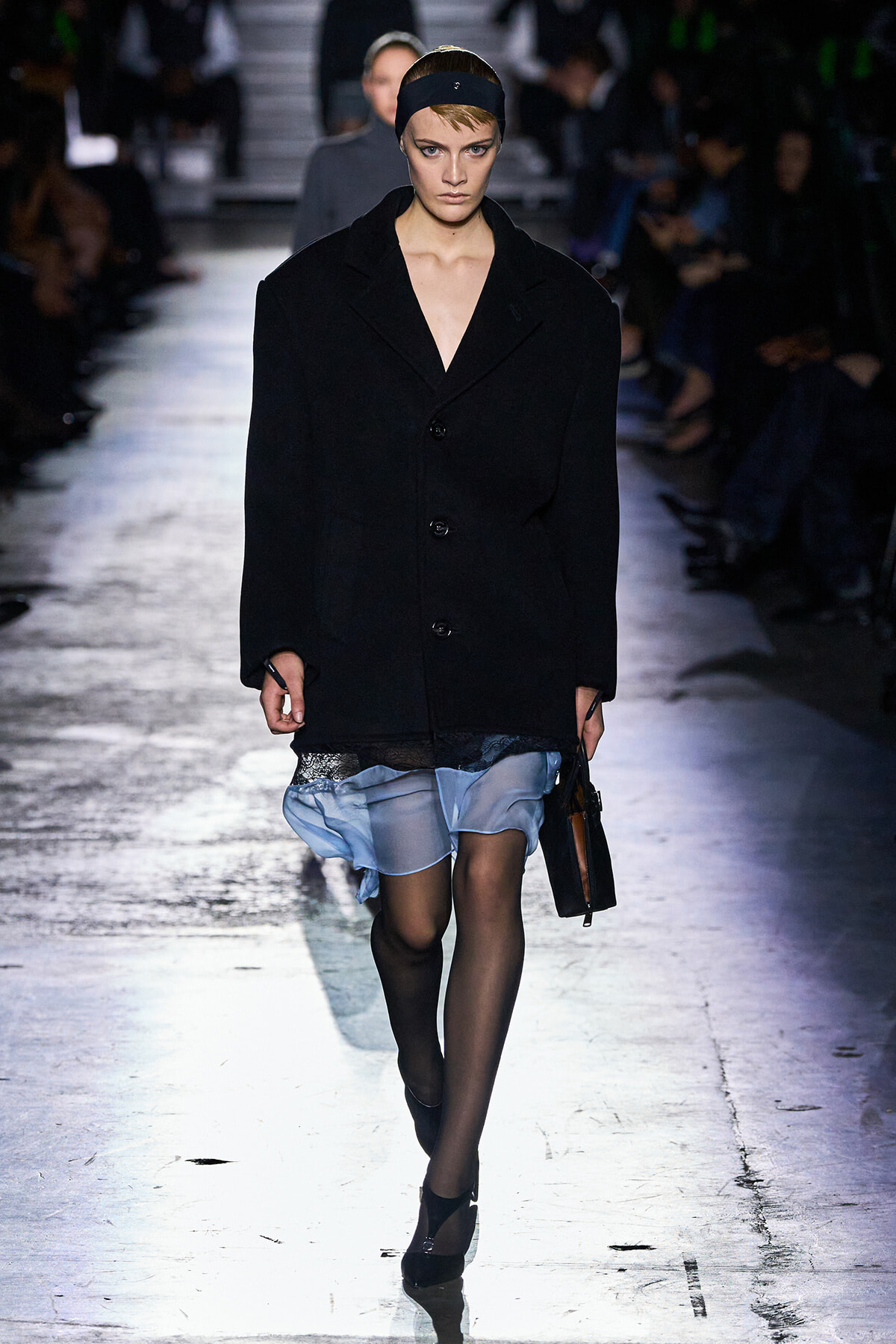 Model walks a fashion show in an oversized black coat over a light blue sheer dress, black tights, headband, and carrying a black handbag on a dim runway.