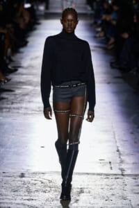 Model on a runway wearing a black turtleneck, dark shorts, and silver studded harness straps with knee-high boots.