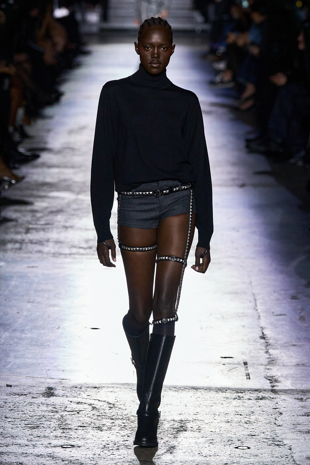 Model on a runway wearing a black turtleneck, dark shorts, and silver studded harness straps with knee-high boots.