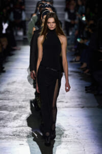Model walking the runway in a black halter top and draped black skirt with a high slit, spotlight on her against a blurred audience backdrop