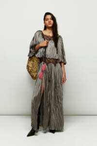 Model wearing a metallic gray pleated gown with black lace trim, standing with a gold textured bag and pink tassels hanging from the strap, against a plain backdrop.