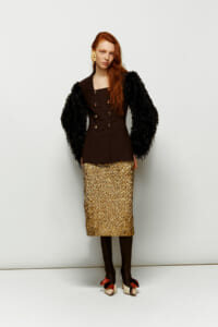 Model wearing a dark brown double-breasted blazer with black feather sleeves and a gold sequined midi skirt, studio pose.