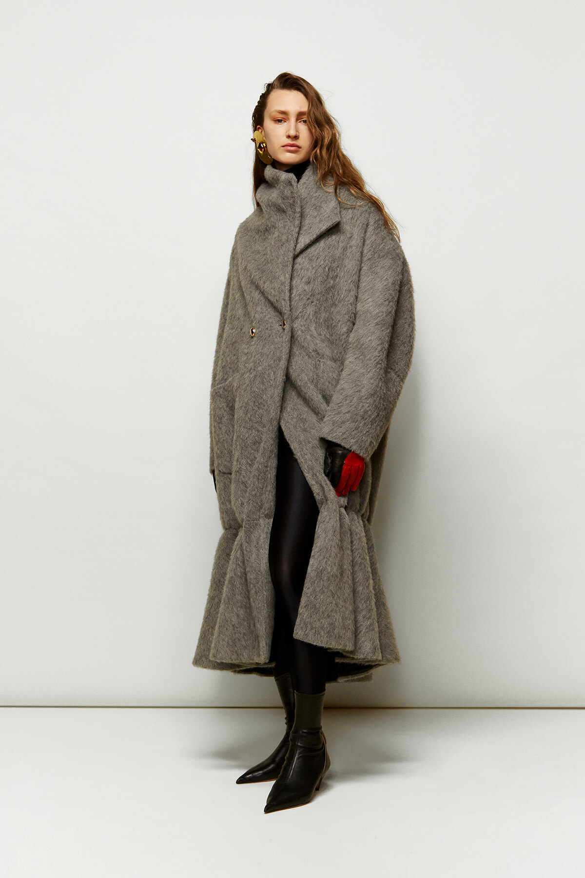Model wearing an oversized gray wool coat with wide lapels, black leggings, and pointed black boots, standing against a light backdrop; left hand in a red glove.