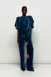 Full-length view of a person wearing a teal velvet draped top and wide-leg pants with a pink tassel belt against a light background.