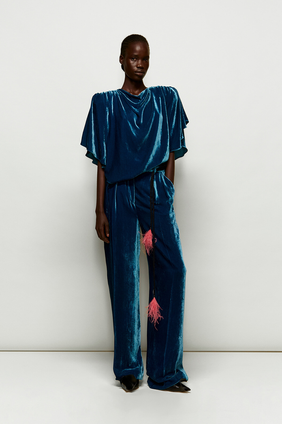 Full-length view of a person wearing a teal velvet draped top and wide-leg pants with a pink tassel belt against a light background.