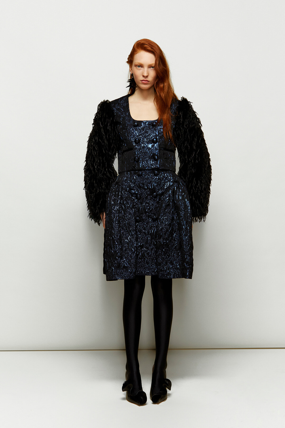 Model wearing a navy textured dress with black feathered sleeves, standing against a light backdrop in tights and heels.