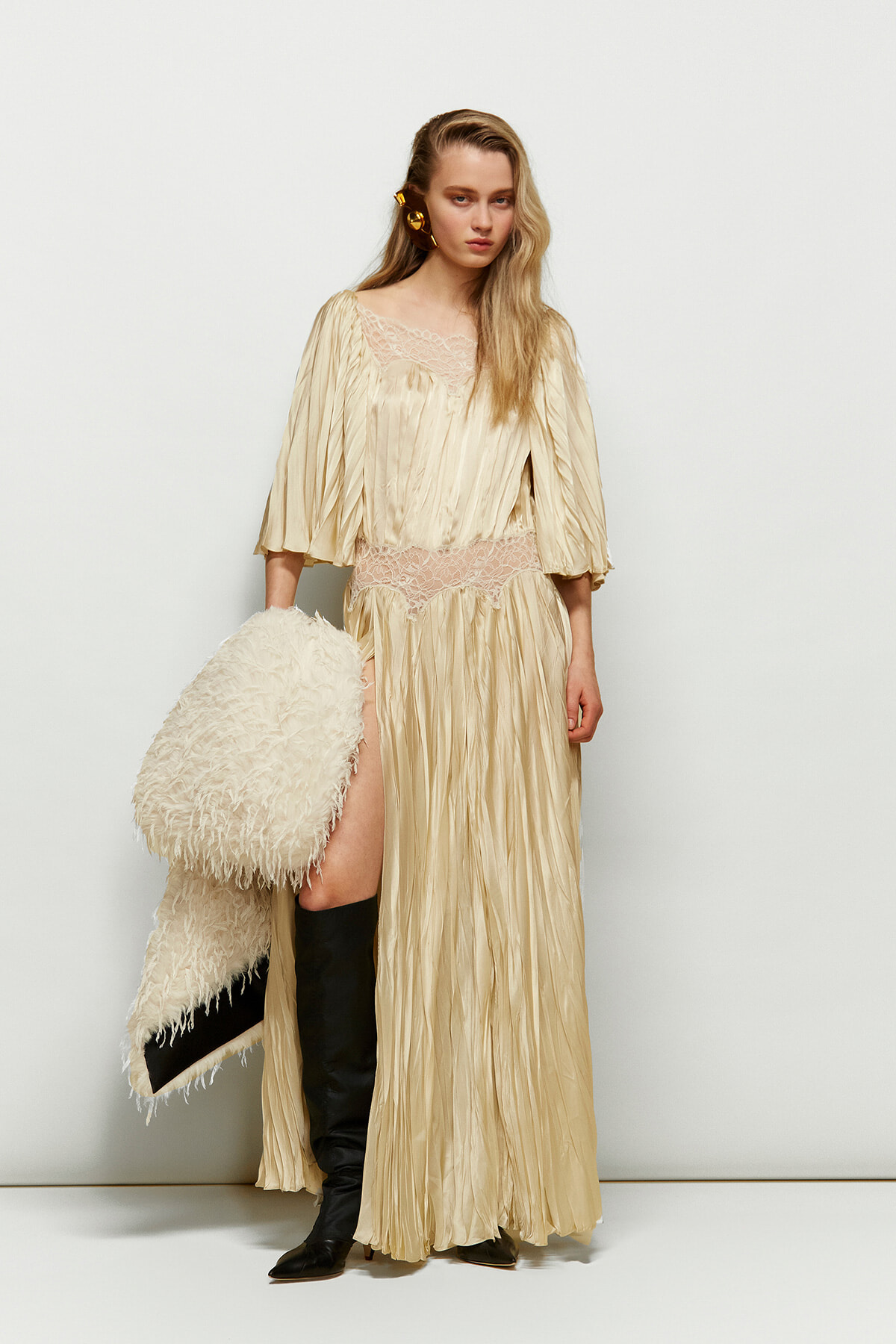 Woman in a champagne-colored pleated gown with a lace bodice, holding a fluffy cream coat and wearing black knee-high boots against a plain backdrop.
