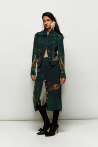 Full-length model wearing a green swan-print coat with matching skirt, gold buttons, black tights, and black heels, accessorized with large white feather earrings against a pale backdrop.
