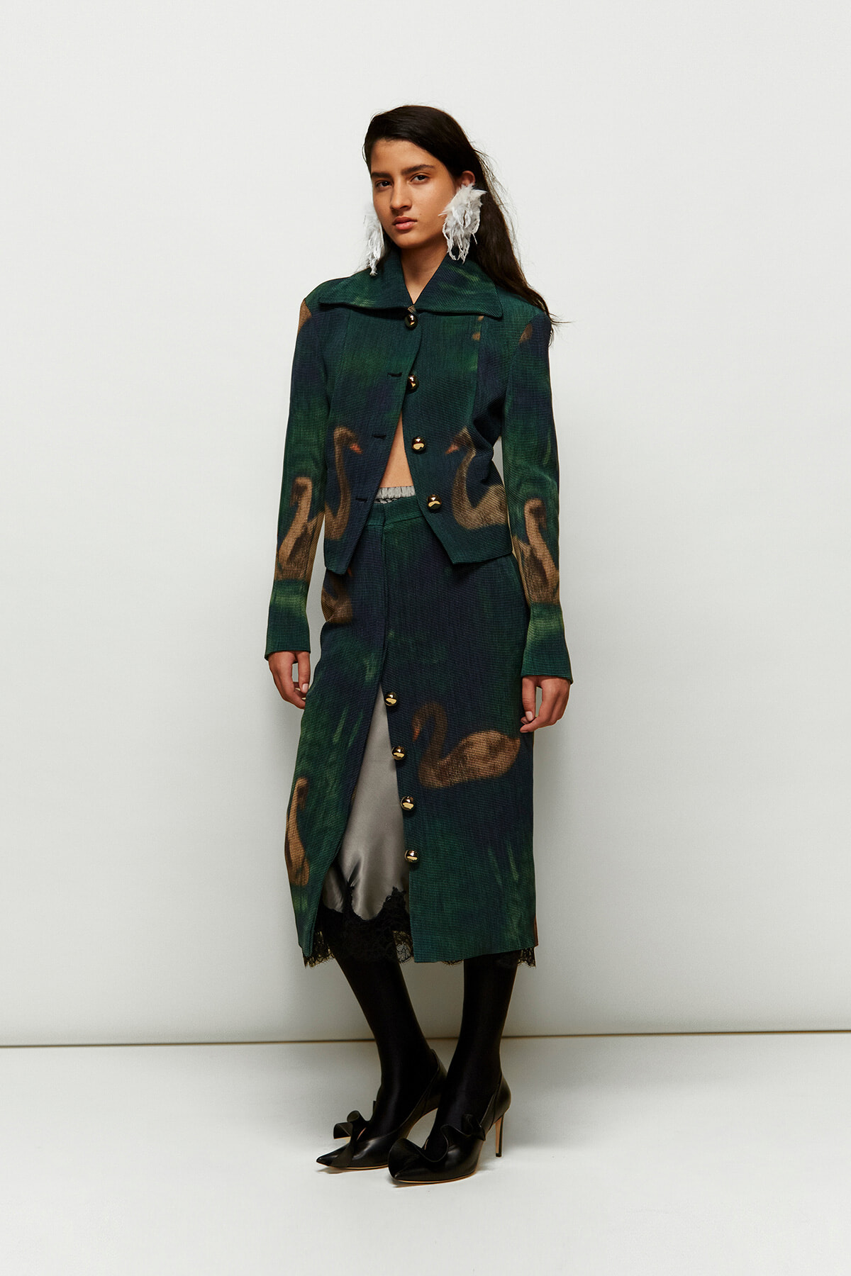 Full-length model wearing a green swan-print coat with matching skirt, gold buttons, black tights, and black heels, accessorized with large white feather earrings against a pale backdrop.