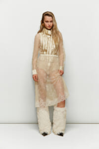 Model in an ivory lace midi dress with sheer sleeves, high neck ruffle, and cream furry boots posed against a plain white backdrop.