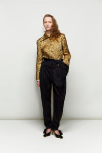 Female model wearing a gold metallic blouse with black pinstripe trousers and a wrapped waist belt, standing against a pale backdrop with black heeled shoes.