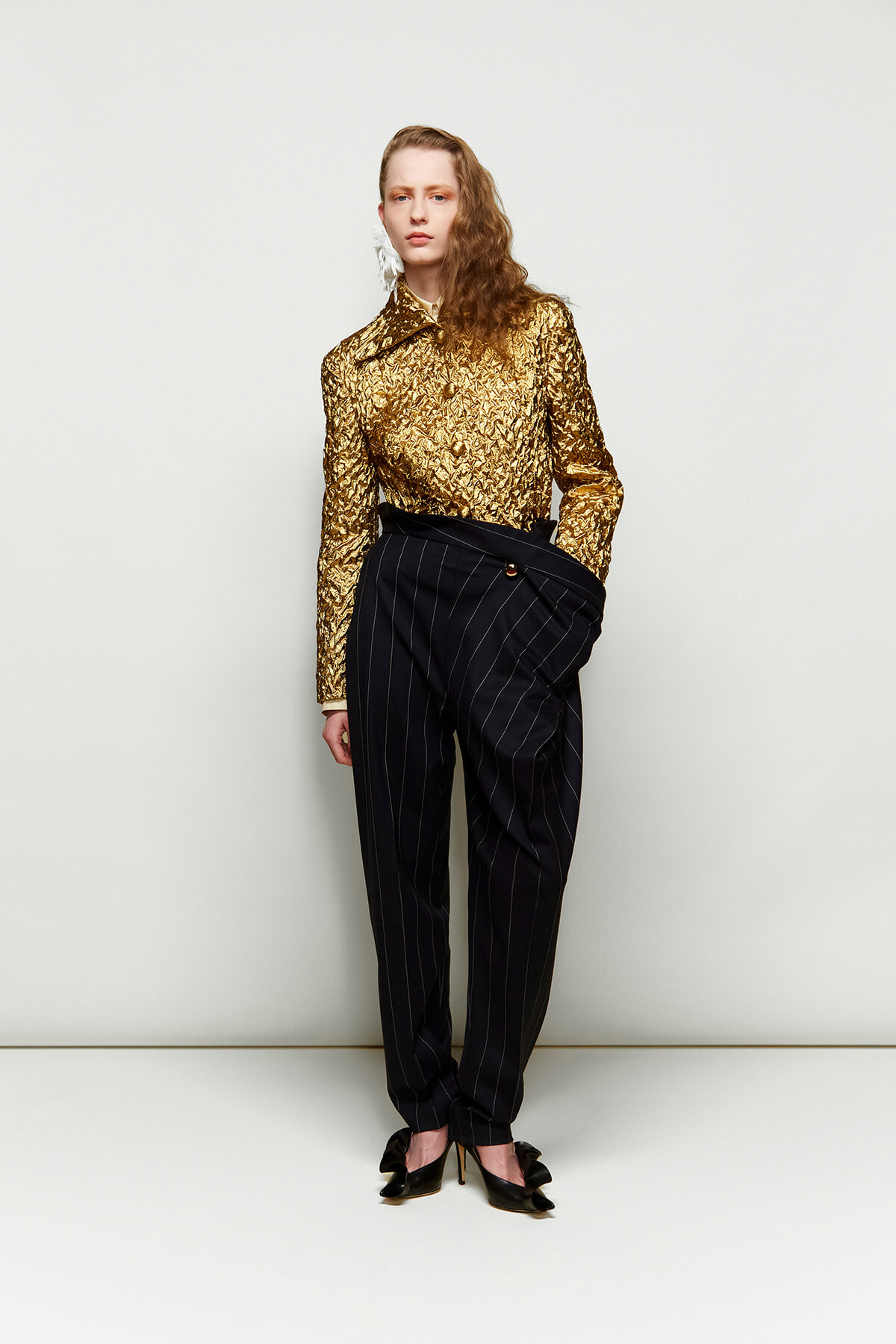 Female model wearing a gold metallic blouse with black pinstripe trousers and a wrapped waist belt, standing against a pale backdrop with black heeled shoes.
